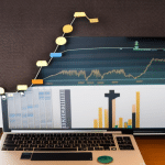 E of a person in front of a laptop with a graph of a rising line and a pile of coins representing different currencies