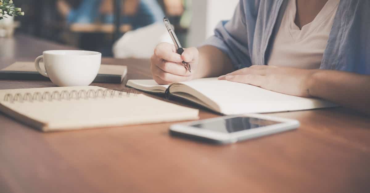 woman-writing-on-a-notebook-beside-teacup-and-tablet-computer